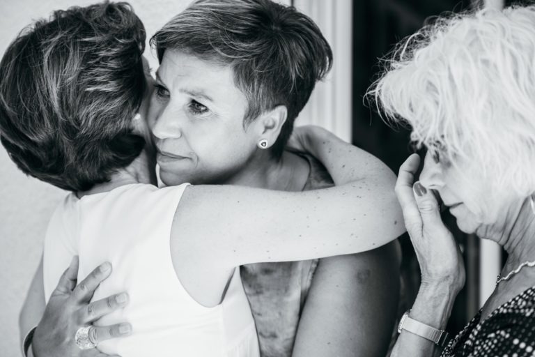 Une photo en noir et blanc montre trois femmes près d'une porte d'entrée à l'intérieur. Deux d'entre elles s'embrassent, l'une fait face à la caméra avec une expression réconfortante. La troisième femme, aux cheveux blancs et aux lunettes, se tient à côté d'elles en essuyant une larme de son œil, évoquant une renaissance émotionnelle.