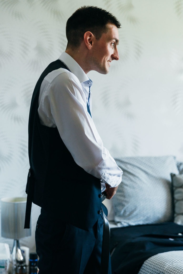 Un homme en chemise blanche et gilet noir se tient dans une chambre à coucher, en train d'ajuster sa cravate. La pièce est dotée d'un mur blanc à motifs, d'un lit avec une couverture grise et blanche et d'une table de chevet avec une lampe blanche et divers petits objets. Avec un air de renaissance émotionnelle, il semble se préparer pour une occasion officielle.