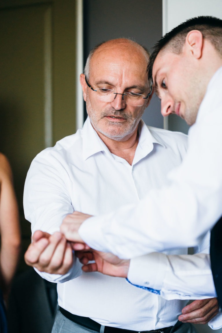 Un homme âgé portant des lunettes ajuste un bouton de manchette pour un homme plus jeune, légèrement penché vers lui. Tous deux sont vêtus de chemises blanches formelles. L'homme âgé semble concentré sur la tâche, tandis que le plus jeune regarde son bouton de manchette, leur connexion évoquant une renaissance émotionnelle tranquille. Une personne partiellement visible se tient à l'arrière-plan.