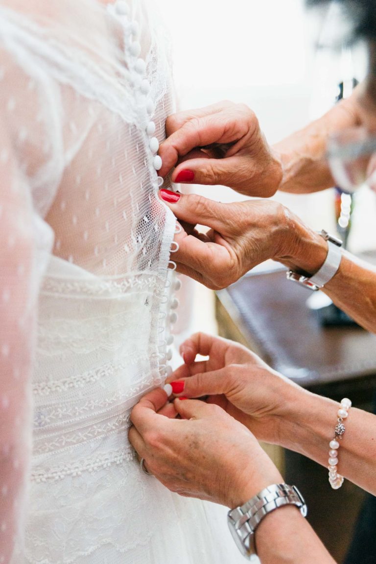 Gros plan de deux paires de mains en train de boutonner le dos de la robe de mariée en dentelle. La mariée est parée d'un délicat corsage en dentelle transparente à pois avec des boutons recouverts de tissu qui descendent jusqu'en bas. Ce moment intime évoque une émotion de renaissance alors qu'une assistante aux ongles peints en rouge et une autre portant une montre l'assistent.