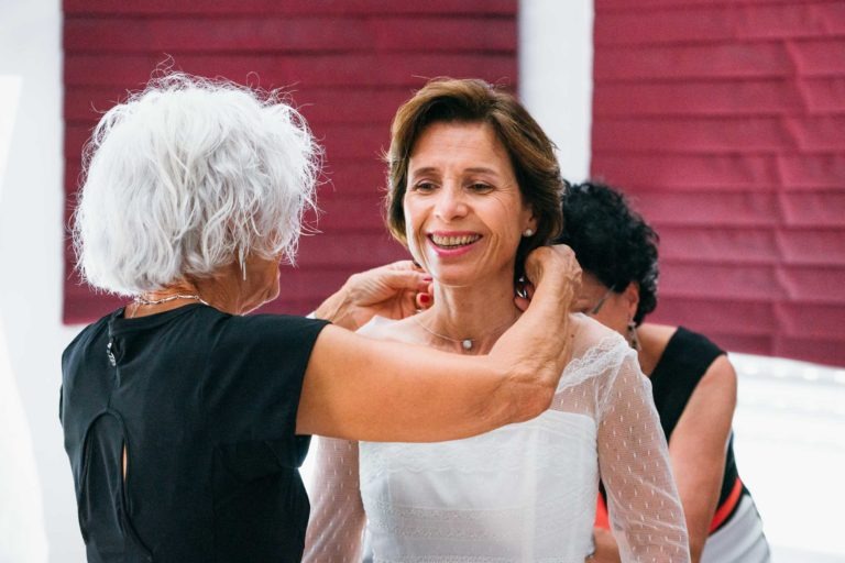 Une femme souriante aux cheveux bruns courts est aidée à enfiler une robe en dentelle blanche par une femme plus âgée aux cheveux blancs courts, évoquant un sentiment de renaissance émotionnelle. Elles sont à l'intérieur, debout devant de grands stores marron. Une autre personne est partiellement visible à l'arrière-plan. L'atmosphère semble joyeuse et chaleureuse.