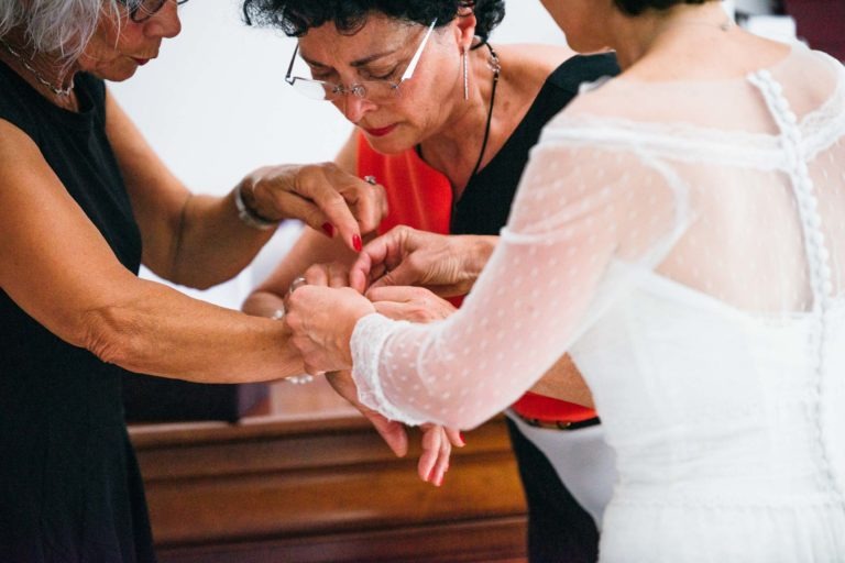 Trois femmes aident à fixer un bracelet au poignet d'une femme portant une robe blanche aux manches transparentes. L'accent est mis sur leurs mains et le bracelet, capturant une émotion de la Renaissance. Une femme a les cheveux courts et bouclés et des lunettes, tandis qu'une autre a les cheveux gris mi-longs et des lunettes.