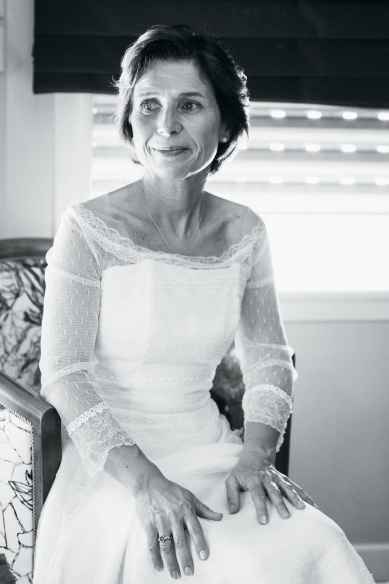 Une femme aux cheveux courts et foncés est assise sur une chaise, vêtue d'une robe de mariée en dentelle à manches longues. Elle a un sourire doux et regarde légèrement sur le côté. La lumière entre par une fenêtre aux stores fermés derrière elle, créant une atmosphère paisible et sereine qui évoque un sentiment de renaissance émotionnelle.