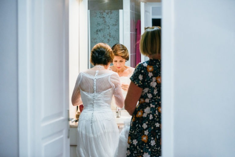 Une mariée en robe de dentelle blanche se tient devant un lavabo de salle de bain, ajustant sa robe devant le miroir. Elle tourne le dos à la caméra. Une autre femme, en robe fleurie, se tient à côté d'elle, face à elle, leur renaissance émotionnelle évidente alors qu'elles se préparent pour la cérémonie de mariage dans une salle de bain lumineuse et moderne.