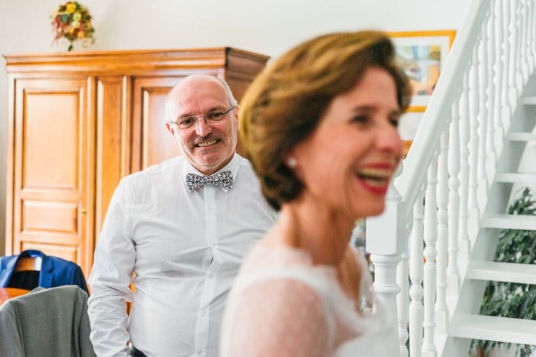 Un homme portant des lunettes et un nœud papillon se tient debout, souriant, à l'arrière-plan, près d'une armoire en bois. Au premier plan, une femme, souriante elle aussi, se détourne de la caméra près d'un escalier blanc. L'environnement suggère un moment joyeux, peut-être festif, une véritable renaissance émotionnelle.