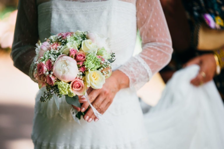 Une mariée en robe de mariée en dentelle blanche tient un bouquet de fleurs composé de roses blanches et roses, de pivoines et de gypsophile. Le bouquet est noué avec un ruban blanc. Embrassant l'atmosphère de renaissance de l'époque, une autre personne se tient derrière la mariée, partiellement visible, aidant à la traîne de la robe.