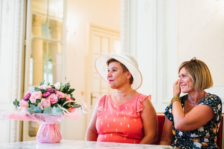 Deux femmes sont assises à une table dans une pièce bien éclairée et décorée avec soin. L'une porte un chapeau blanc et un haut sans manches corail à motifs floraux, l'autre porte une robe fleurie à manches courtes. Un bouquet de fleurs roses est posé sur la table devant elles. La femme en corail sourit, tandis que l'autre semble vivre une renaissance émotionnelle.