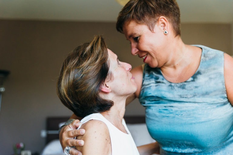 Deux femmes partagent un moment de tendresse, debout près l'une de l'autre et se souriant chaleureusement. Toutes deux ont les cheveux courts ; l'une porte un haut blanc sans manches tandis que l'autre arbore un haut sans manches à motifs bleu et gris. L'une des femmes tient doucement l'épaule de l'autre, dégageant affection et connexion dans leur renaissance émotionnelle.