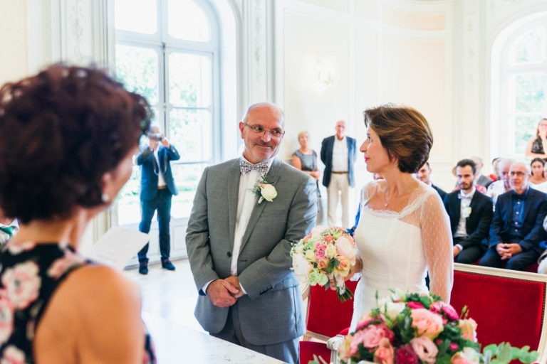 Dans une salle lumineuse aux grandes fenêtres, un couple de mariés se tient face à face, en présence de nombreux invités. Le marié, vêtu d'un costume gris et d'un nœud papillon, tient un bouquet. La mariée, vêtue d'une robe blanche, tient également un bouquet. Une femme en robe fleurie, peut-être une officiante, se tient à proximité tandis qu'ils vivent leur renaissance émotionnelle.