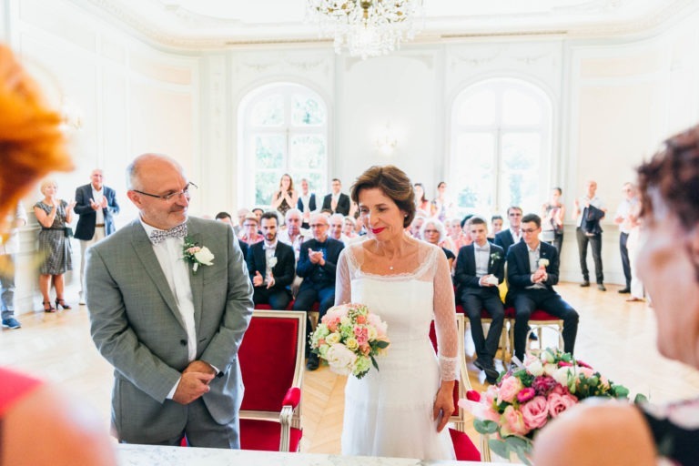 Une mariée et un marié se tiennent devant une salle lumineuse et élégante remplie d'invités qui applaudissent. Le marié, en costume gris clair et nœud papillon, regarde la mariée. La mariée porte une robe blanche et tient un bouquet. La salle est dotée de grandes fenêtres, de lustres et d'un décor blanc ornementé, évoquant une atmosphère de renaissance.