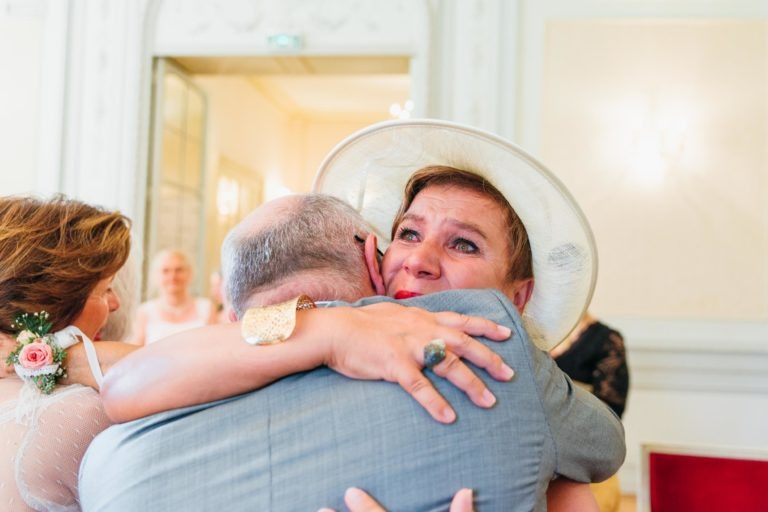 Une femme portant un chapeau blanc et des boucles d'oreilles en forme de larme serre dans ses bras un homme en costume gris, son visage exprimant l'émotion avec des larmes dans les yeux - une aura de renaissance émotionnelle. Elle porte une bague et un bracelet. Une femme avec une boutonnière rose est partiellement visible à gauche. Le décor semble être une pièce élégante et lumineuse.