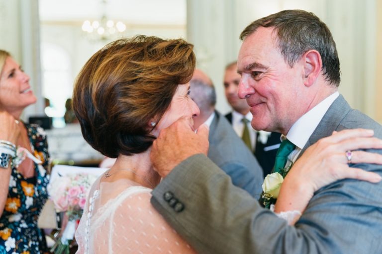 Un couple de personnes âgées en tenue de soirée, l'homme en costume gris et cravate verte et la femme en robe blanche, partagent un moment de tendresse lors d'une réunion. L'homme tient doucement la joue de la femme, tous deux souriant chaleureusement, incarnant une renaissance émotionnelle. Derrière eux, d'autres personnes, dont une femme en robe fleurie, engagent la conversation.