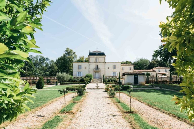 Une villa pittoresque à deux étages avec une façade beige et un toit noir se dresse au bout d'un chemin de gravier flanqué de rangées symétriques de haies taillées et de petits arbres. La verdure luxuriante du jardin encadre la scène, capturant une sensation émotionnelle de renaissance, tandis qu'un ciel lumineux au-dessus arbore une légère traînée de condensation en diagonale.