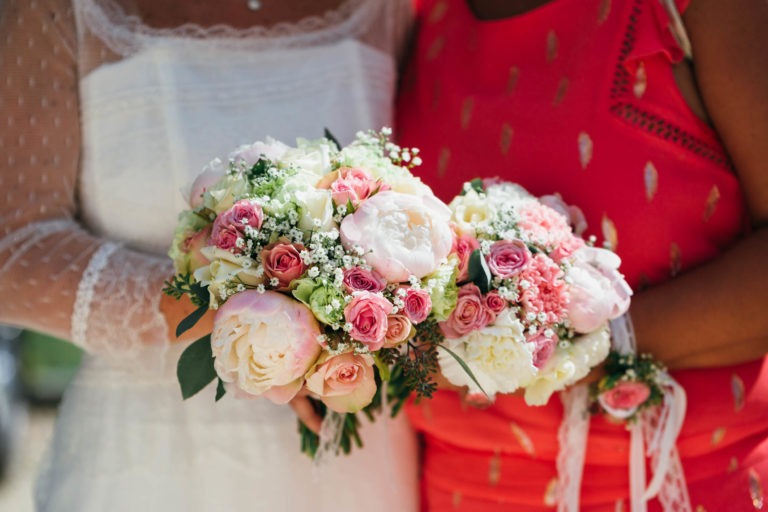 Gros plan de deux personnes tenant des bouquets de fleurs aux couleurs vives, évoquant une atmosphère de renaissance. Les bouquets sont composés de roses roses et blanches, de pivoines et de verdure. Une personne porte une robe en dentelle blanche et l'autre une robe rouge à motifs dorés. Leurs têtes ne sont pas représentées, l'accent étant mis sur les fleurs et la tenue.