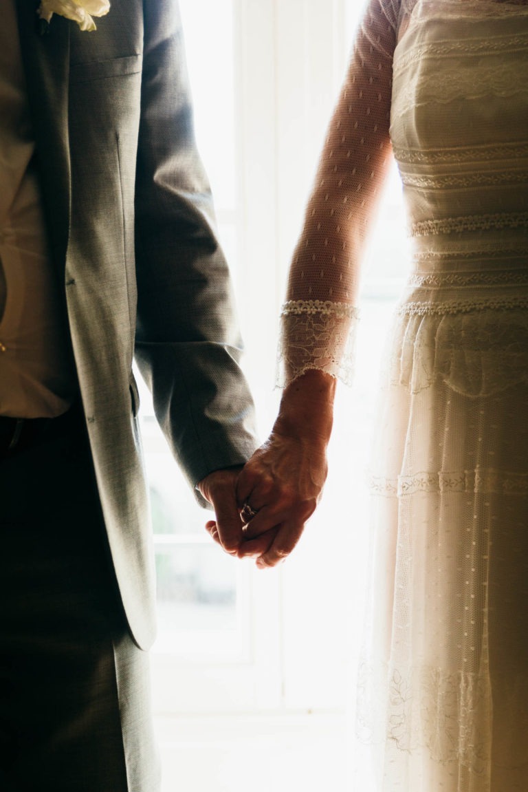 Un couple habillé en tenue de soirée se tient par la main. La personne à gauche porte un costume gris avec une chemise blanche, tandis que la personne à droite porte une robe blanche à manches longues en dentelle délicate. Ils se tiennent près d'une fenêtre bien éclairée, baignée d'une douce lumière du soleil, capturant leur renaissance émotionnelle.