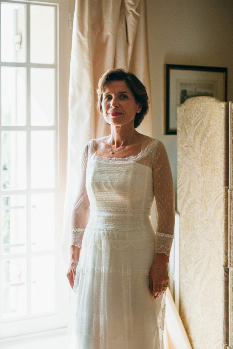 Une femme vêtue d'une robe blanche en dentelle à manches longues se tient à l'intérieur, près d'une fenêtre lumineuse avec des rideaux blancs. Elle a les cheveux courts et foncés et affiche une expression calme. À l'arrière-plan, il y a un paravent avec des motifs ornementaux et des œuvres d'art encadrées sur le mur. La lumière remplit la pièce, créant une atmosphère sereine évoquant la Renaissance émotionnelle.