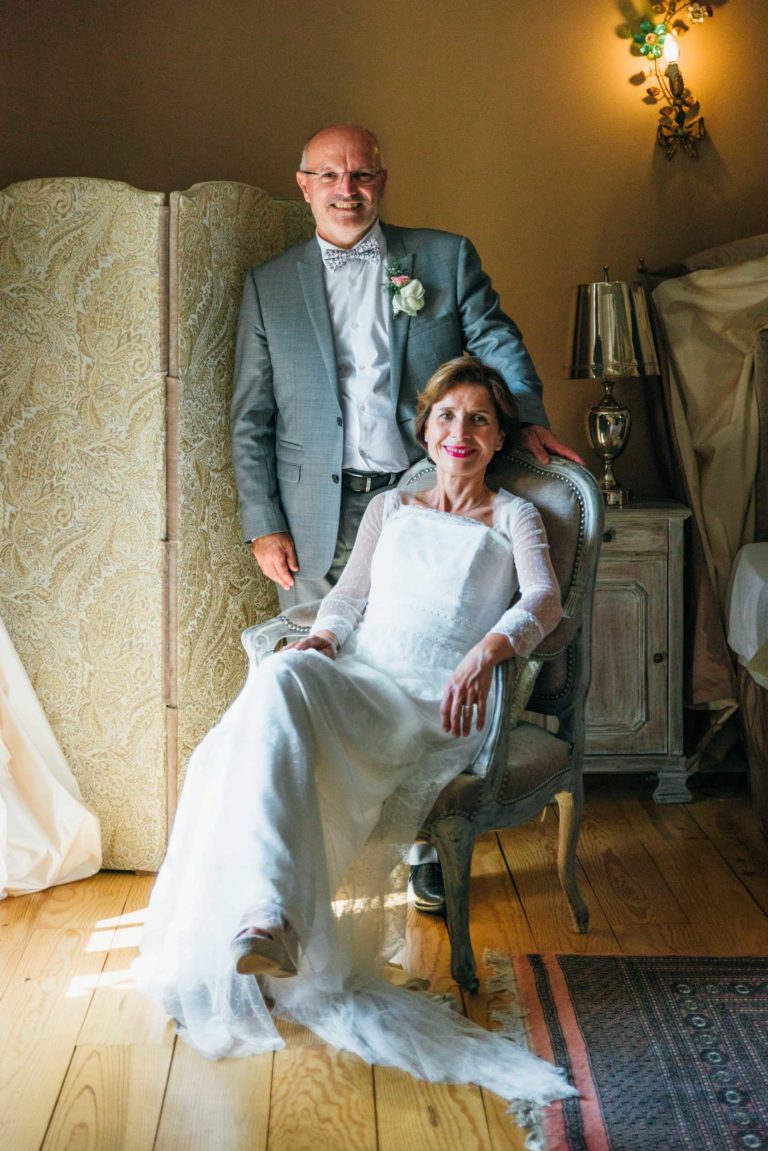 Un couple pose à l'intérieur le jour de leur mariage, dégageant une émotion de renaissance. Le marié, en costume gris et nœud papillon, se tient derrière la mariée assise dans une robe blanche aux manches en dentelle. Ils se trouvent dans une pièce de style vintage aux tons beiges, avec un parquet en bois et un paravent décoratif.