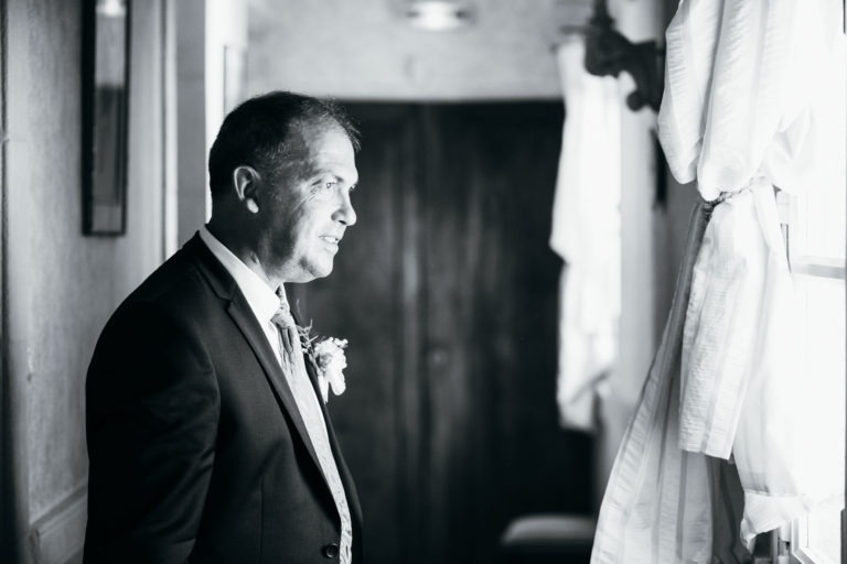 Un homme vêtu d'un costume sombre avec une boutonnière se tient à l'intérieur, près d'une fenêtre à rideau, regardant dehors avec une expression pensive. La photo, rendue en noir et blanc, évoque une renaissance émotionnelle, créant une atmosphère intemporelle et contemplative. Une porte floue persiste en arrière-plan.