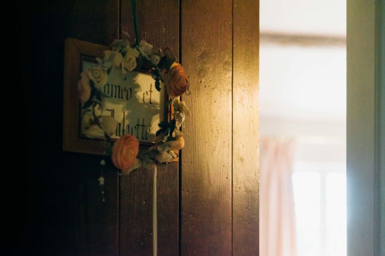 Une porte en bois ornée d'une couronne de fleurs suspendue entourant les mots « home et labette » écrits à l'intérieur d'un cadre. La lumière d'une pièce adjacente illumine doucement la scène, révélant une porte partiellement ouverte avec un aperçu d'une pièce lumineuse dotée d'une fenêtre ensoleillée avec des rideaux lumineux - une émotion de renaissance.