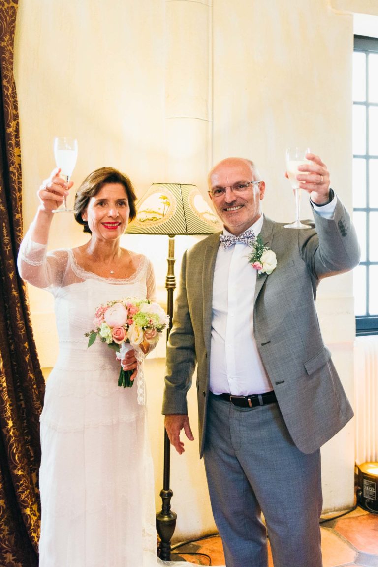 Un couple souriant, habillé en tenue de soirée, lève ses verres de champagne pour porter un toast, exprimant une atmosphère de renaissance. La femme, vêtue d'une robe de mariée blanche, tient un bouquet de fleurs. L'homme porte un costume gris avec un nœud papillon et une boutonnière. Ils se tiennent à côté d'une lampe ornée d'un abat-jour beige, sur un fond chaleureusement éclairé.