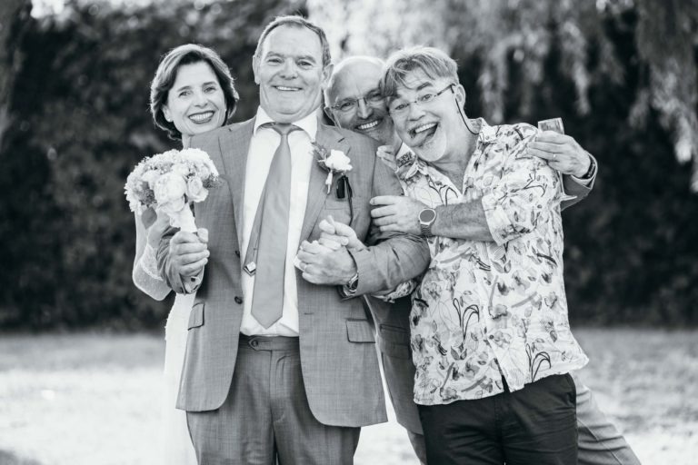 Une photo en noir et blanc montre une scène joyeuse lors d'un mariage. Deux personnes vêtues de leurs tenues de mariage, tenant des fleurs, se tiennent au centre, souriantes. Deux autres, l'une portant une chemise imprimée, les enlacent des deux côtés, tous souriant largement à l'objectif. L'arrière-plan est un jardin flou débordant d'émotions de la Renaissance.