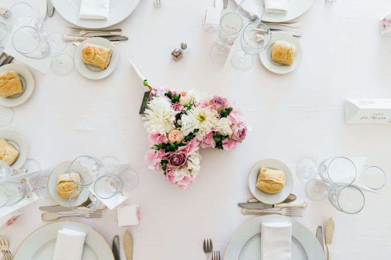 Une table à manger bien dressée comprend des assiettes blanches avec des petits pains, des verres en cristal et des couverts en argent sur une nappe blanche impeccable. Un bouquet de fleurs, dont des fleurs blanches, roses et violettes, sert de pièce maîtresse. De petits marque-places et des cartes de menu sont soigneusement disposés autour de la table, évoquant une atmosphère de renaissance.