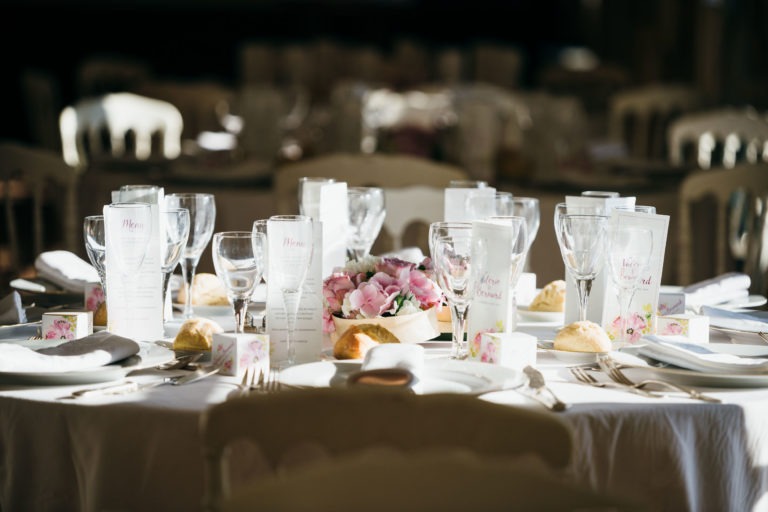 Une table ronde joliment dressée lors d'un événement officiel, illuminée par la lumière du soleil. La table est composée de nappes blanches, de verres soigneusement disposés, de couverts et d'assiettes avec des petits pains. Au centre, il y a une composition florale avec des fleurs roses. Des menus et des marque-places sont disposés autour de la table, capturant une ambiance émotionnelle de la Renaissance.