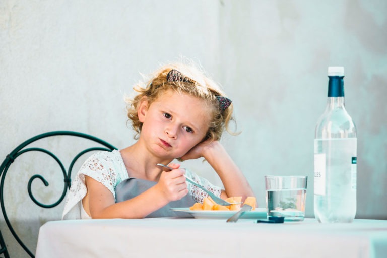 Une jeune fille aux cheveux blonds bouclés est assise à une table blanche. Elle pose sa tête sur une main, évoquant une atmosphère de renaissance, et tient une fourchette dans l'autre, à côté d'une assiette avec de la nourriture. Sur la table se trouvent un verre d'eau et une bouteille transparente. Elle porte une robe blanche et un bandeau, avec une chaise en fer forgé derrière elle.