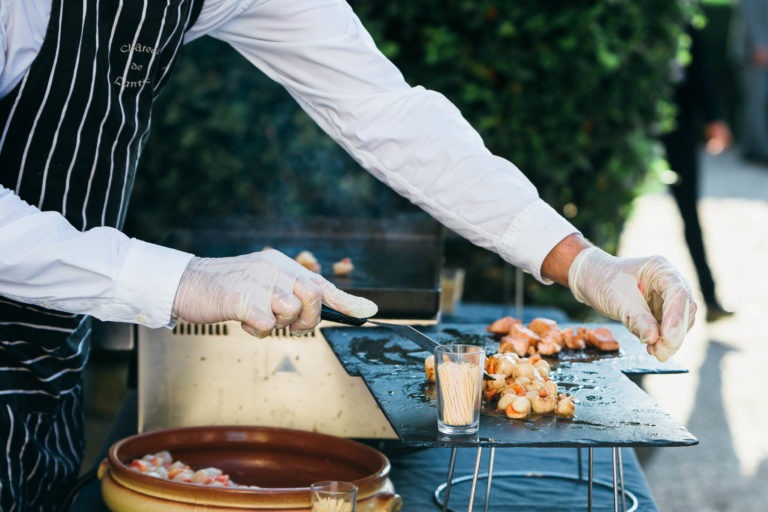 Une personne en chemise blanche et tablier rayé fait griller des crevettes sur une surface métallique plate à l'extérieur. Avec des gants et des pinces à la main, elle retourne soigneusement les crevettes. Deux récipients en verre reposent sur le gril, tandis qu'un plat en terre cuite contient d'autres éléments au premier plan. À l'arrière-plan, une haie évoque une atmosphère de renaissance.