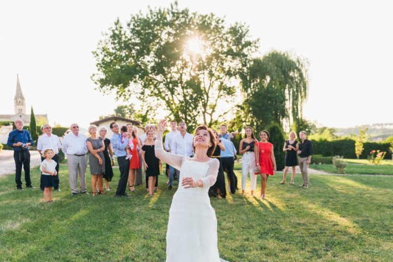 Une mariée en robe blanche se tient dans un espace extérieur herbeux, se préparant à lancer son bouquet. Derrière elle, un groupe d'invités au mariage attendent avec impatience de l'attraper. La scène, pleine d'émotions de la Renaissance, est éclairée par le soleil couchant avec un grand arbre et de la verdure en arrière-plan, créant un cadre pittoresque.