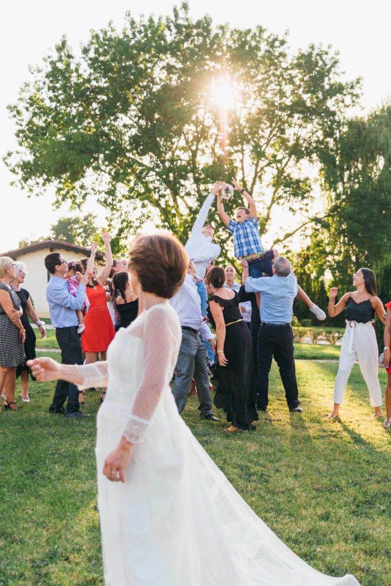 Une mariée en robe blanche à manches longues se tient au premier plan, tandis qu'un groupe de personnes en tenue décontractée et semi-formelle fait la fête à l'arrière-plan. Un homme soulève un enfant dans les airs, entouré d'arbres et de soleil filtrant à travers les branches, créant une scène de renaissance émotionnelle.