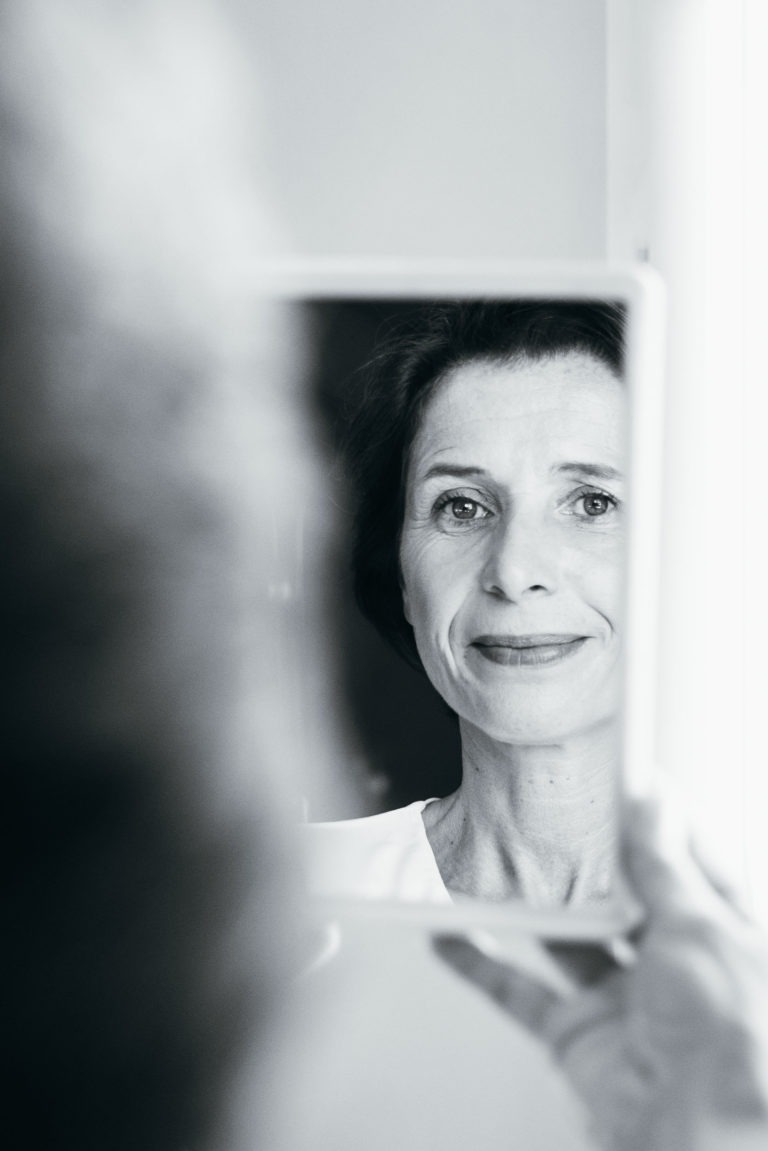 Une photographie en noir et blanc montre une femme d'âge moyen aux cheveux noirs courts qui regarde son reflet dans un petit miroir. Le miroir est tenu près de son visage, capturant de manière vivante son expression calme et légèrement souriante. L'arrière-plan est flou, soulignant l'émotion de renaissance sur son visage serein.