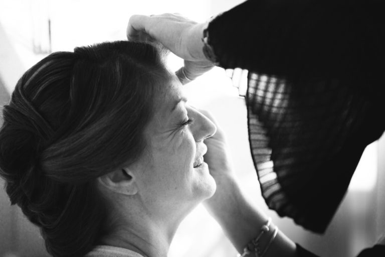 Une photo en noir et blanc montrant un gros plan de profil d'une femme souriante, les yeux fermés, tandis que la main d'une autre personne ajuste doucement ses cheveux. Les cheveux de la femme sont soigneusement coiffés et elle semble détendue, capturant le moment tendre d'une surprise de mariage. La deuxième personne n'est visible que par son bras et sa main, portant une manche sombre et fluide.