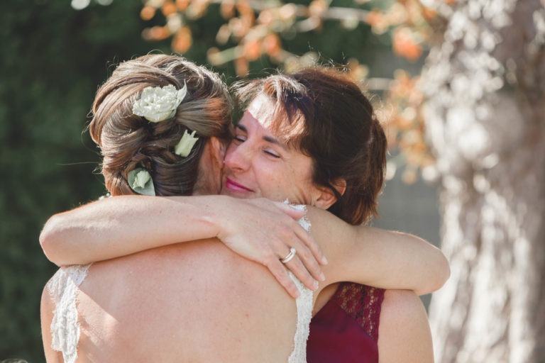 Une femme aux cheveux tressés et vêtue d'un vêtement clair enlace une autre femme vêtue d'une robe marron. Elles s'embrassent chaleureusement à l'extérieur, entourées de feuillage vert et de fleurs orange. C'est un moment de surprise de mariage, car la femme en robe marron sourit les yeux fermés.