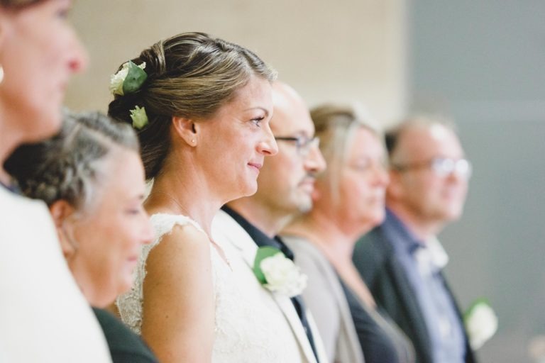 Une mariée souriante, vêtue d'une robe en dentelle blanche avec des fleurs dans les cheveux, se tient en rang avec d'autres personnes, toutes tournées dans la même direction. L'arrière-plan est flou, attirant l'attention sur leurs profils latéraux. Le groupe semble faire partie d'une cérémonie de mariage surprise.