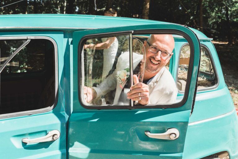 Un homme en costume blanc et lunettes sourit à travers la portière ouverte d'une voiture vintage bleu sarcelle, tenant une petite boutonnière florale. L'arrière-plan montre une silhouette floue dans une robe blanche et un feuillage vert, faisant allusion à une surprise de mariage en plein air. L'atmosphère semble festive et joyeuse.