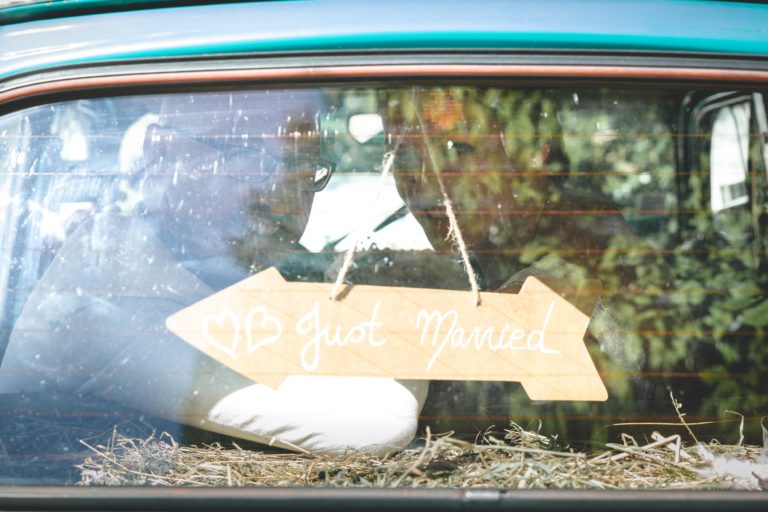 Une photo prise à travers la lunette arrière d'un véhicule montre un couple assis à l'intérieur, avec un panneau marron clair en forme de flèche sur lequel est écrit « Just Married » et un petit symbole de cœur sur la gauche. Une touche de foin est visible à l'intérieur, ajoutant à la surprise de leur mariage.