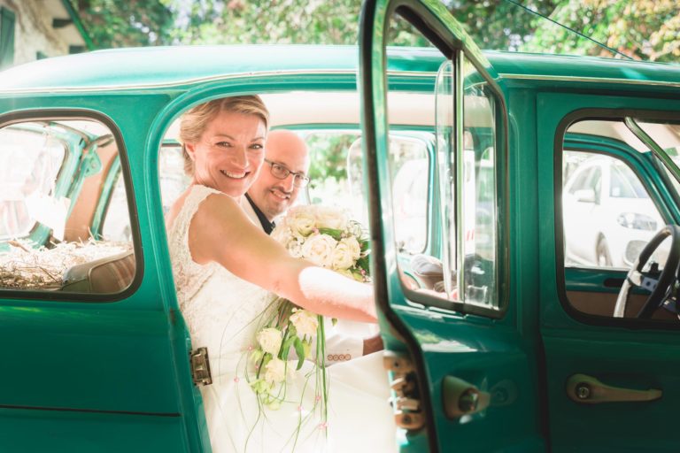 Un couple joyeux le jour de leur mariage, vêtu d'une tenue de soirée, sourit alors qu'ils sont assis dans une voiture turquoise vintage. La mariée, en robe blanche, tient un bouquet de fleurs de couleur claire de leur surprise de mariage, tandis que le marié, en costume, est partiellement visible derrière elle. La portière de la voiture est ouverte, révélant une journée ensoleillée à l'extérieur.