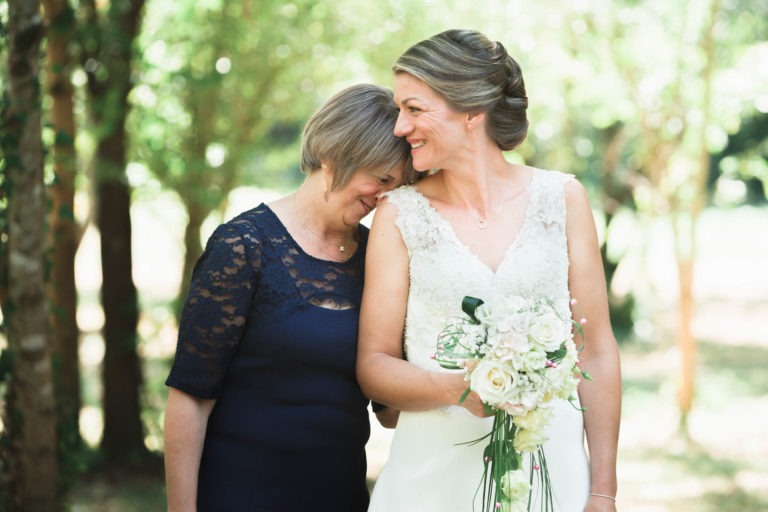 Une mariée en robe de dentelle blanche et tenant un bouquet de fleurs blanches et rose pâle se tient à l'extérieur avec une femme en robe de dentelle bleu marine. La femme plus âgée appuie affectueusement sa tête sur l'épaule de la mariée, toutes deux souriant chaleureusement, capturant l'essence de leur joyeuse surprise de mariage dans la zone boisée ensoleillée.