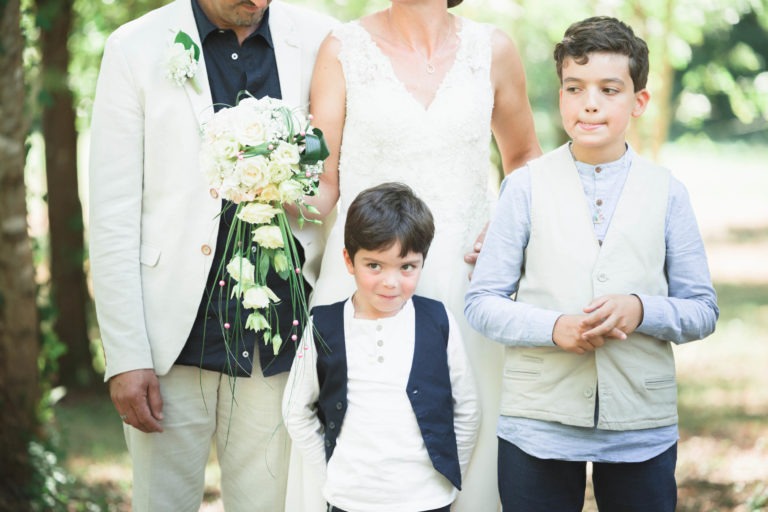 Une photo de famille dans un jardin montre quatre personnes. Un homme adulte en costume clair et une femme adulte en robe blanche tiennent un bouquet de fleurs blanches, faisant allusion à une possible surprise de mariage. Devant eux se tiennent deux garçons, tous deux en chemise blanche et gilet foncé. Le plus jeune regarde l'objectif avec un sourire timide.