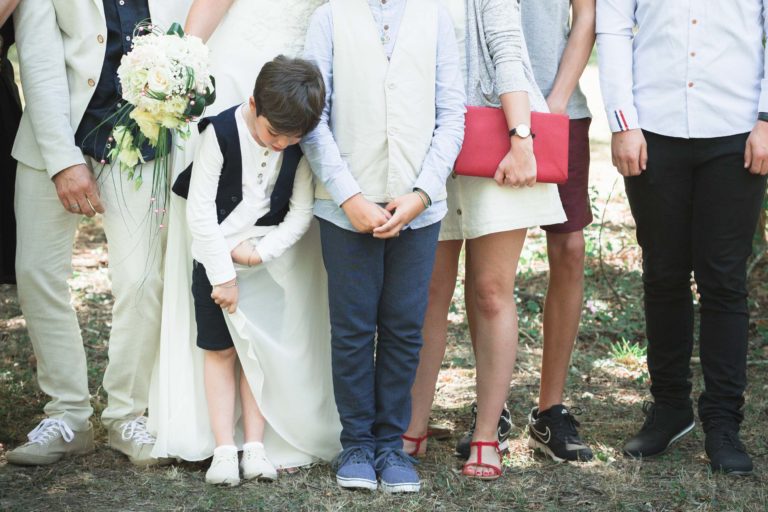 Un groupe de personnes se tient dehors, seules leurs moitiés inférieures étant visibles. Un adulte en robe blanche tient un bouquet de fleurs blanches, suggérant une surprise de mariage. Un jeune garçon en gilet et short regarde vers le bas, touchant la robe. Un autre enfant se tient à côté de lui. D'autres sont habillés en tenue décontractée et semi-formelle.