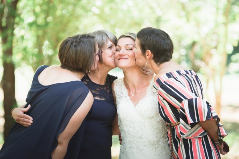 Quatre femmes se trouvent dans un décor extérieur avec une végétation luxuriante en arrière-plan. Les femmes de gauche et de droite embrassent les joues de la femme souriante au centre, qui porte une robe de mariée blanche. C'est une joyeuse surprise de mariage, avec ses amies habillées respectivement de tenues sombres et rayées.