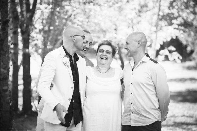 Une photo en noir et blanc capture un moment de joie lors d'un mariage surprise avec quatre personnes debout à l'extérieur, souriant et riant ensemble. Deux hommes sur le côté gauche, l'un portant un blazer clair et l'autre une chemise foncée, une femme au milieu portant une robe blanche et un homme à droite portant une chemise claire. Des arbres et de la verdure sont visibles en arrière-plan.