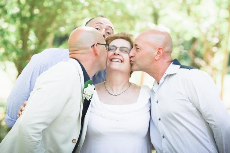 Une femme âgée pleine de joie est embrassée avec amour sur les deux joues par deux hommes chauves, chacun tenant ses épaules. Un troisième homme se tient derrière eux, se penchant de manière enjouée et souriant à la caméra. Ils sont dehors au milieu d'une verdure et d'arbres lumineux, portant des vêtements de couleur claire, célébrant peut-être une surprise de mariage.