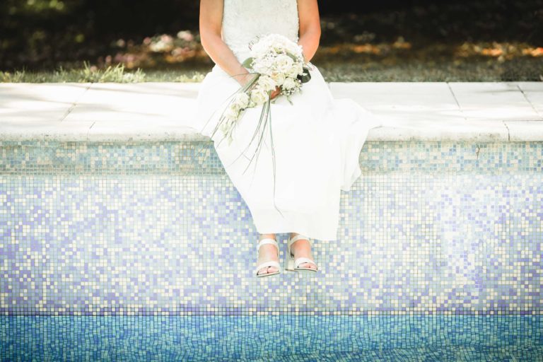 Une personne vêtue d'une tenue blanche est assise au bord d'une piscine carrelée et vide. Ses jambes pendent sur le bord et elle porte des sandales blanches. Elle tient sur ses genoux un bouquet de fleurs blanches aux tiges vertes, donnant à la scène un air de surprise de mariage. Les carreaux de la piscine sont un mélange de bleu et de blanc, et l'arrière-plan est flou.