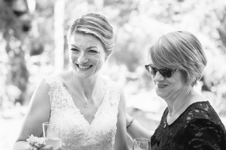 Photo en noir et blanc de deux femmes souriantes en plein air. La femme de gauche porte une robe de mariée en dentelle, tient un verre et a les cheveux relevés. La femme de droite porte des lunettes de soleil, une robe en dentelle sombre et a les cheveux courts. Elles semblent profiter d'un moment de bonheur ensemble lors d'un mariage surprise.