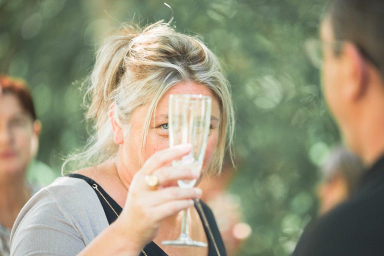 Une femme aux cheveux clairs, partiellement attachée, tient un verre transparent pour porter un toast lors de ce qui semble être une surprise de mariage. Elle semble regarder la personne devant elle. L'arrière-plan est flouté par de la verdure, ce qui indique un décor extérieur. Plusieurs autres personnes sont faiblement visibles à l'arrière-plan.