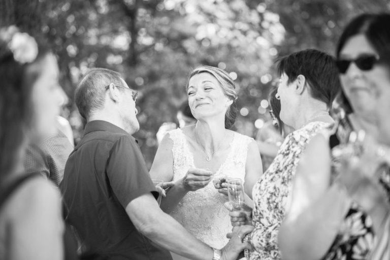 Une photo en noir et blanc montre une mariée en robe blanche, debout à l'extérieur avec un sourire doux, tenant un verre. Elle est entourée d'invités, tous apparemment engagés dans une conversation lors de ce qui semble être une surprise de mariage. Les arbres et les feuilles sont flous en arrière-plan, indiquant un cadre extérieur avec lumière naturelle.