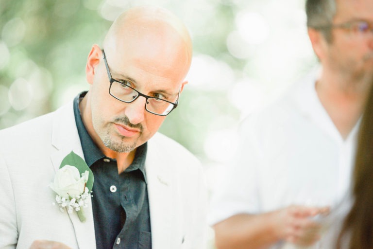 Un homme chauve avec des lunettes et une barbe claire porte une veste de costume claire avec une boutonnière de rose blanche, parfaite pour une surprise de mariage. Il regarde légèrement vers la gauche avec une expression pensive. L'arrière-plan est doucement éclairé et flou, avec une autre personne partiellement visible sur la droite.