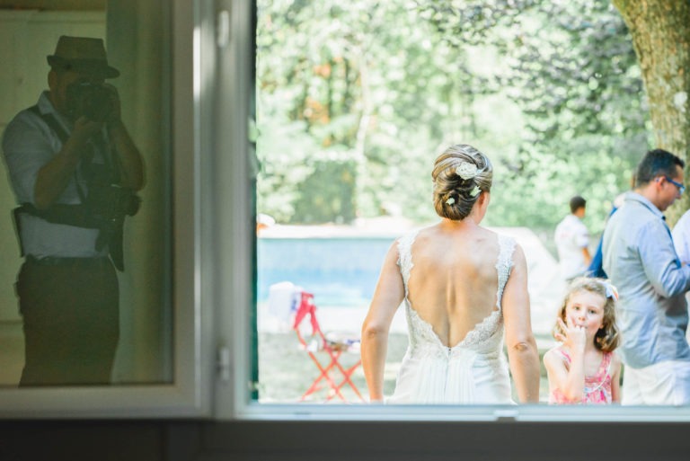 Une mariée dans une robe blanche dos nu avec des détails en dentelle se tient près d'un arbre dans un cadre extérieur ensoleillé, le dos tourné vers l'extérieur. Une jeune fille en robe rose se tient à proximité. À travers une grande fenêtre, un photographe capture la joyeuse surprise du mariage. D'autres invités sont vus en train de célébrer en arrière-plan.